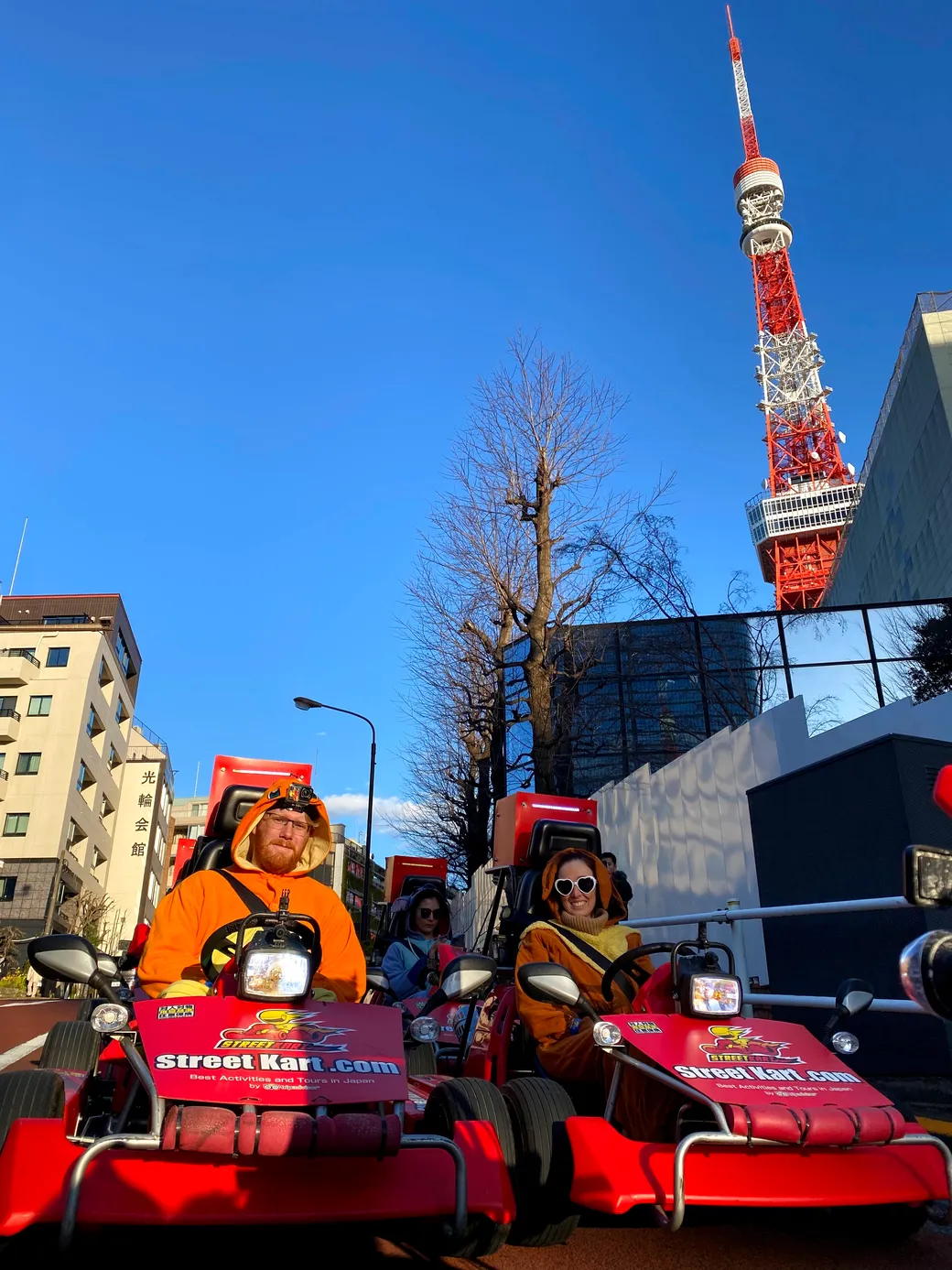 Two people in orange outfits drive red Street Kart go-karts on a city street, with Tokyo Tower in the blue sky behind them.