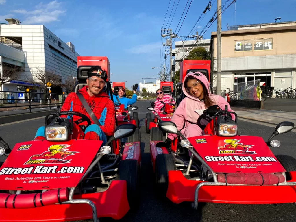 People in red go-karts driving on a city street, smiling and waving at the camera; kart fronts display StreetKart.com logo and text.