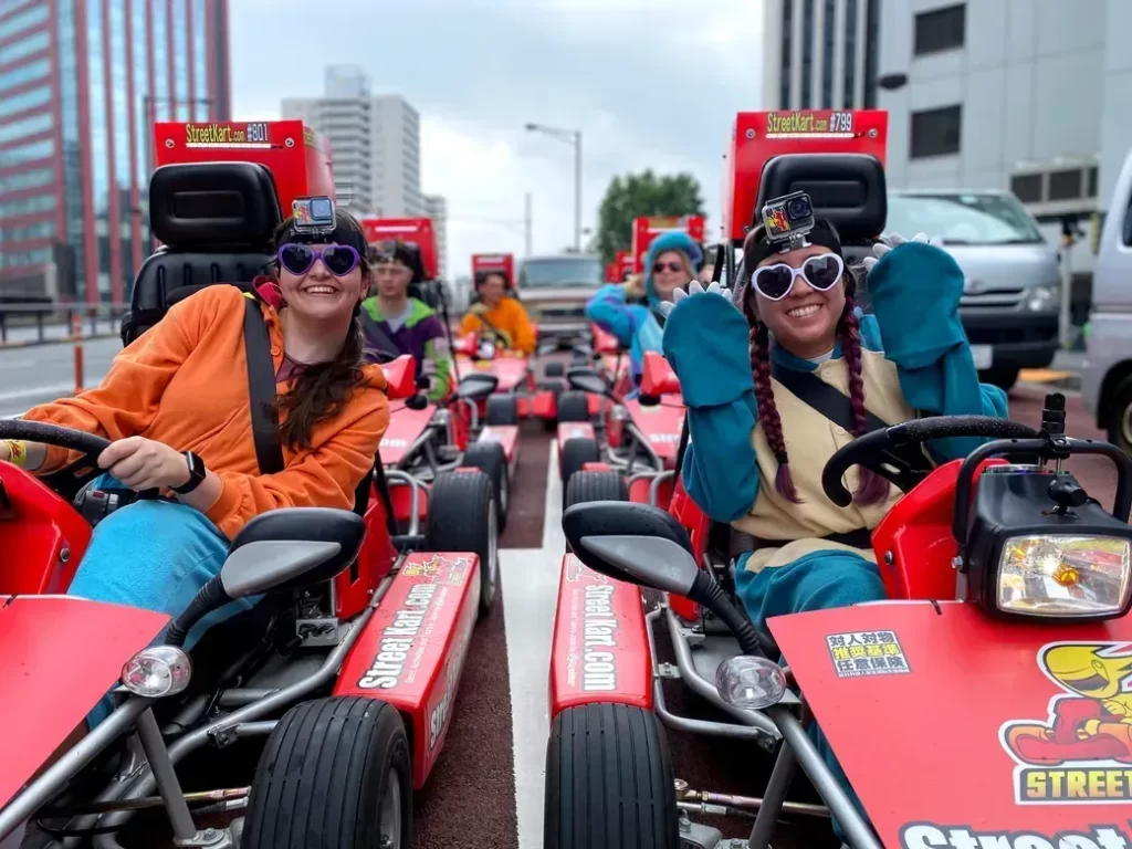 Two women in red StreetKart go-karts smile at the camera, wearing heart-shaped sunglasses and GoPro cameras on their helmets.